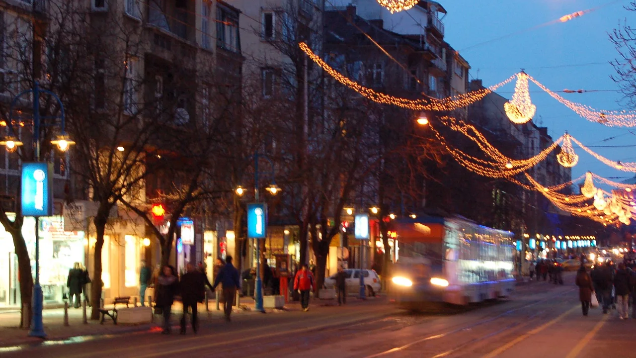 Strings of festive lights and lit chandeliers hanging above a central Sofia street at dusk, with a tram coming towards the camera