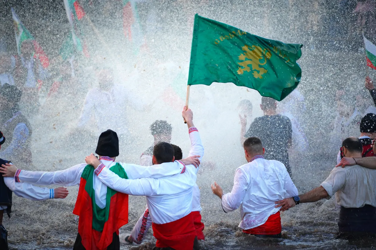Men in white embroidered shirts standing waist-deep in a winter river with linked arms, one holding a green flag, spray flying around them