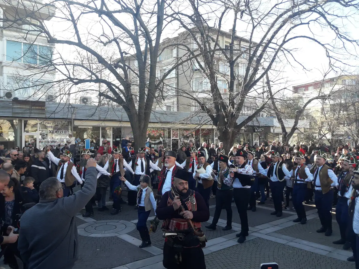 A large group of men in white shirts, waistcoats and feathered caps dancing in a Bulgarian town square while a piper in costume plays at the front