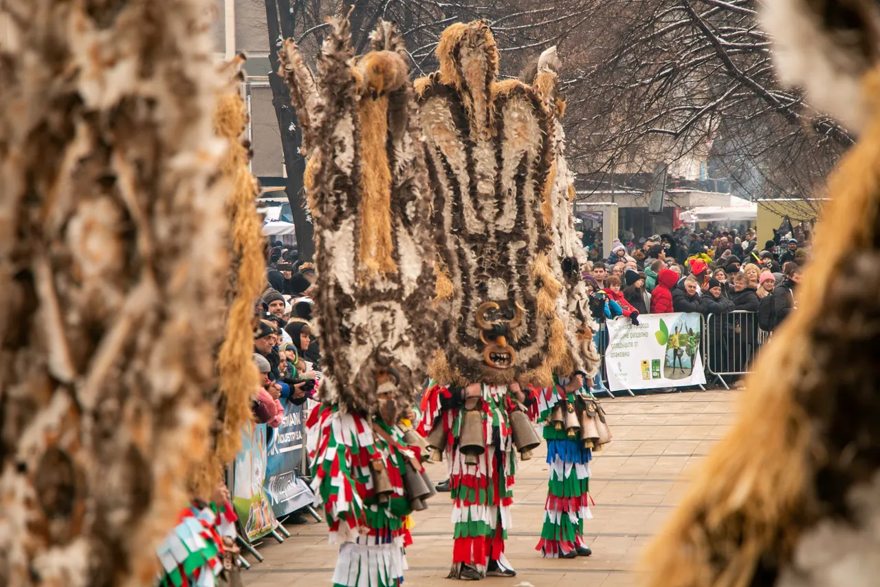 Kukeri in towering fur masks and red-green-white fringed costumes hung with heavy bronze bells parading past crowds behind barriers