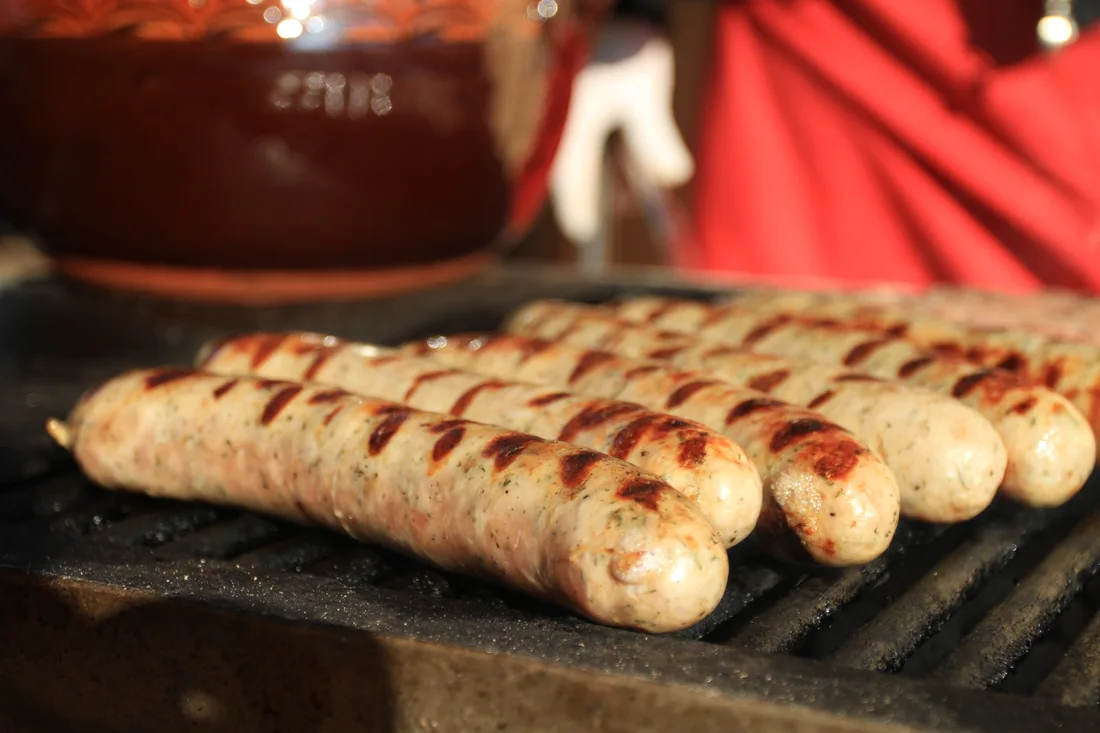 Bulgarian pork sausages browning on a grill at a Christmas market stall, with a clay pot of mulled wine behind