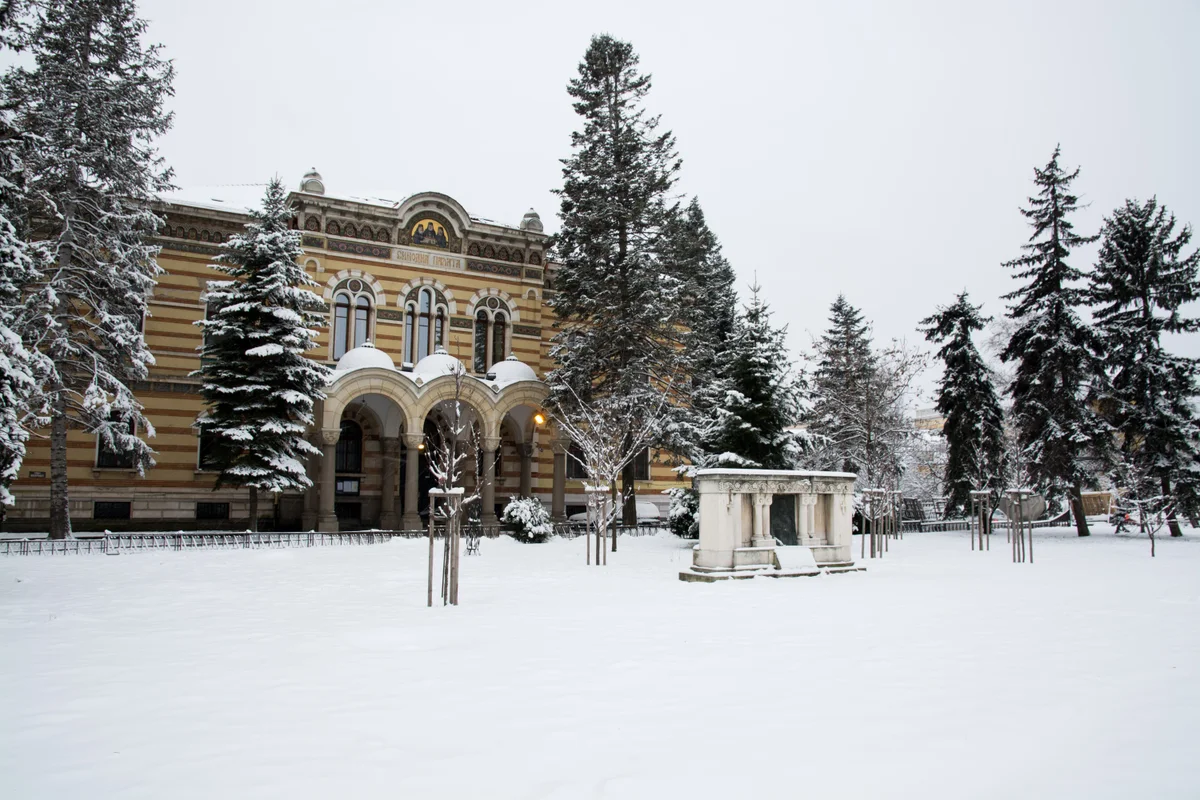 Deep untouched snow across a park in central Sofia with snow-laden conifers and a striped nineteenth-century building behind
