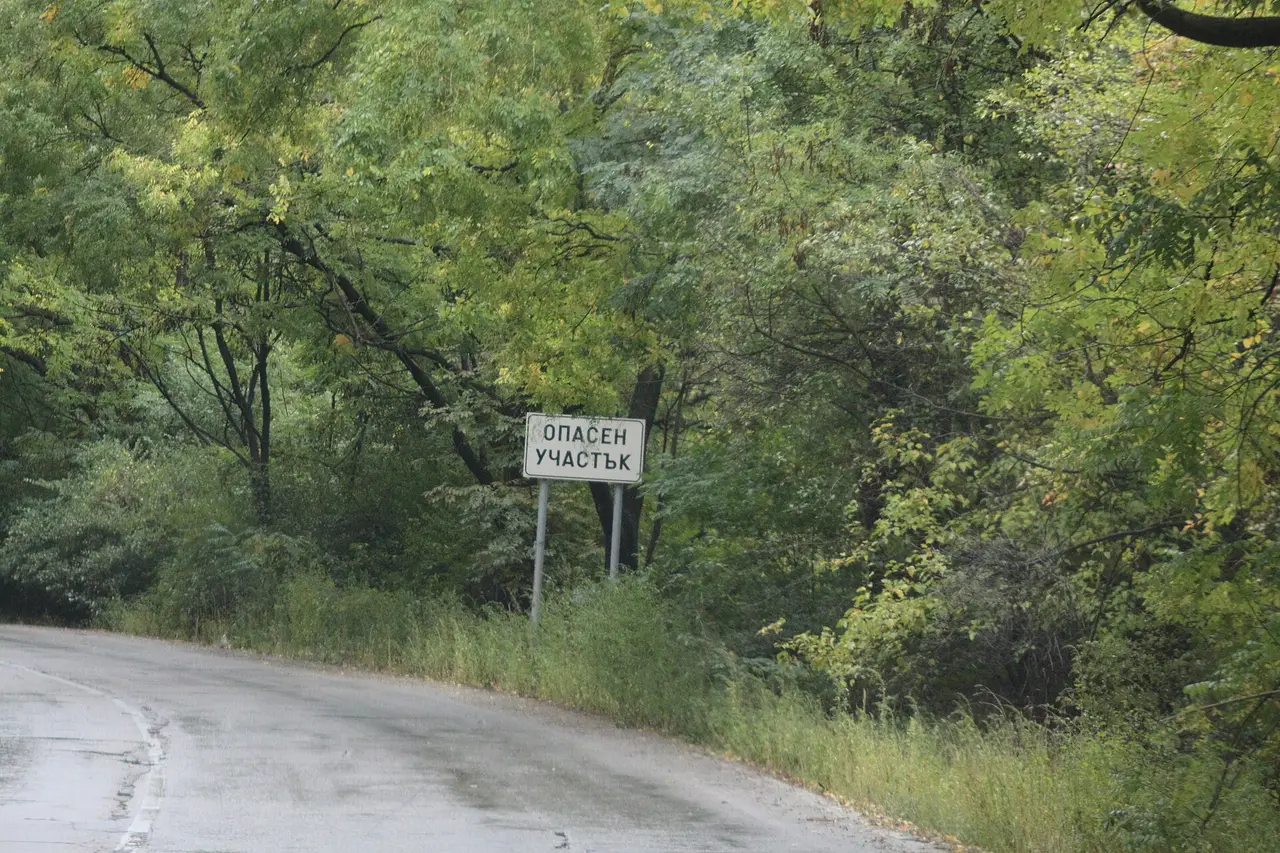 A red-bordered warning road sign on a rural Bulgarian road marking a dangerous stretch, with hills behind