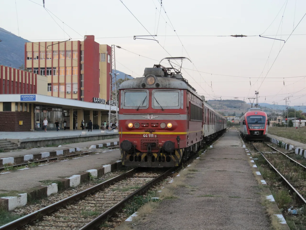 A red BDZ locomotive-hauled passenger train at Karlovo station with a modern Desiro railcar on the next platform and mountains behind