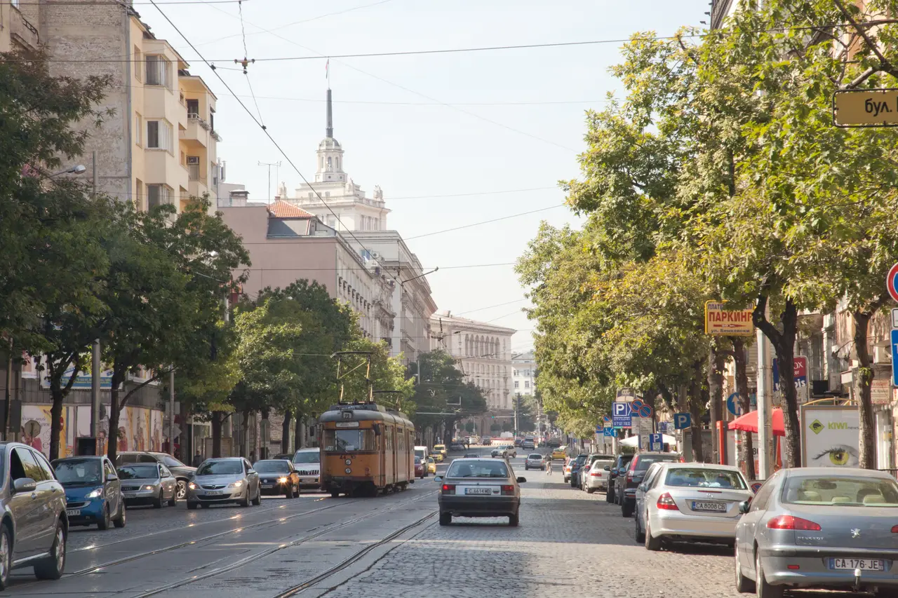 An orange tram running down a tree-lined boulevard in central Sofia among cars, with the tower of the former Party House behind