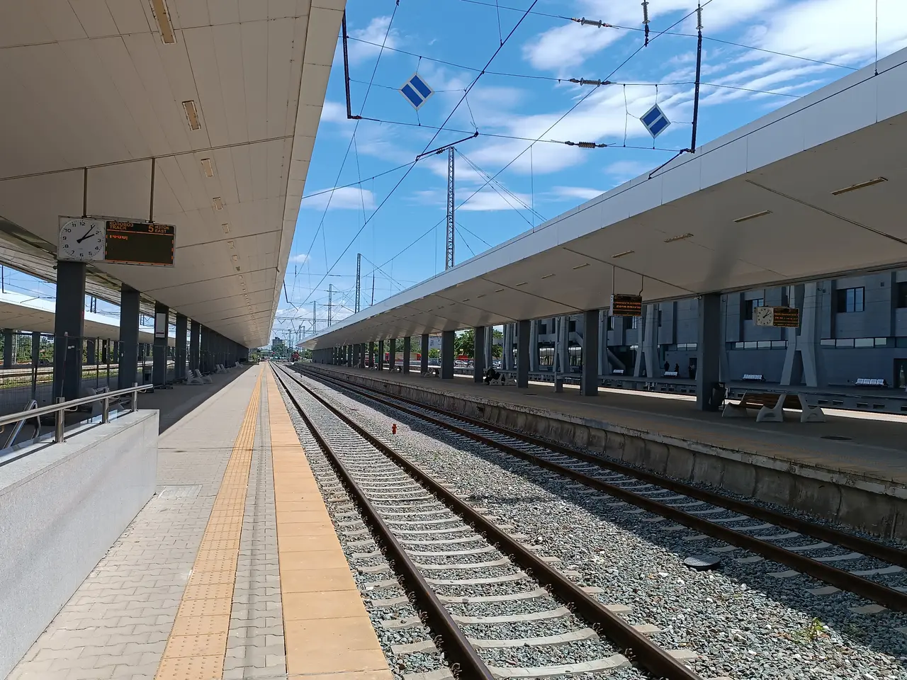 The clean, covered platforms of Sofia Central Station under a blue sky, with tracks, overhead wires and a departures clock