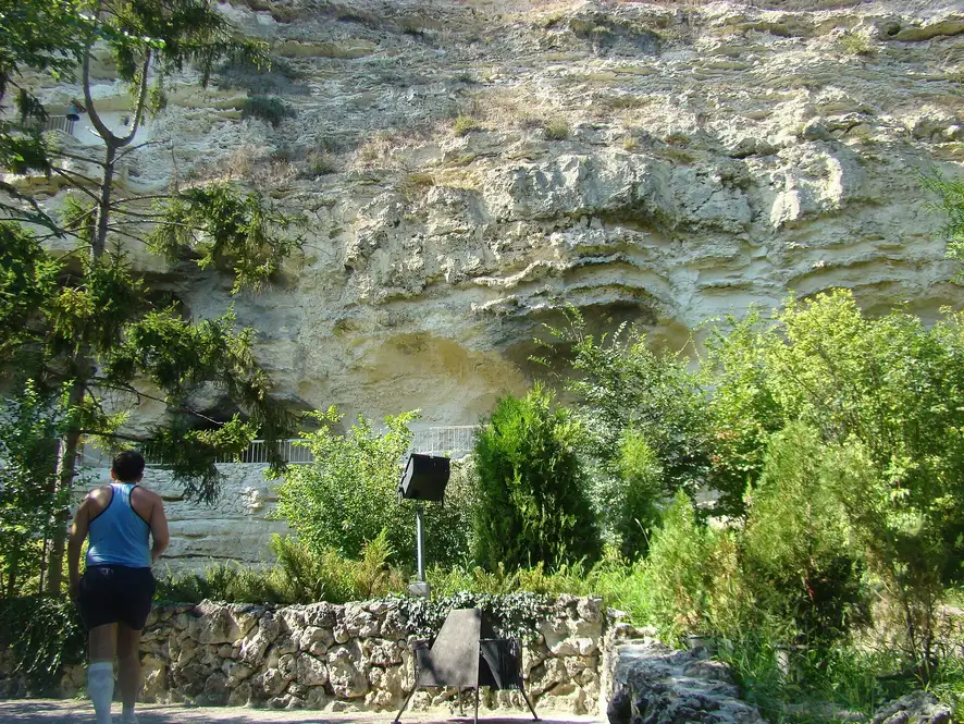 The Aladzha rock monastery, cave chambers and openings cut into a tall pale limestone cliff face, with green trees and a low stone wall in front