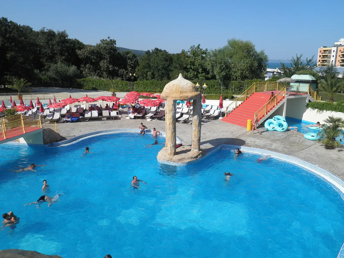 The Aquapolis water park at Golden Sands, a blue pool with a stone parasol fountain, red sunbeds and water slides, the forested hills and sea behind