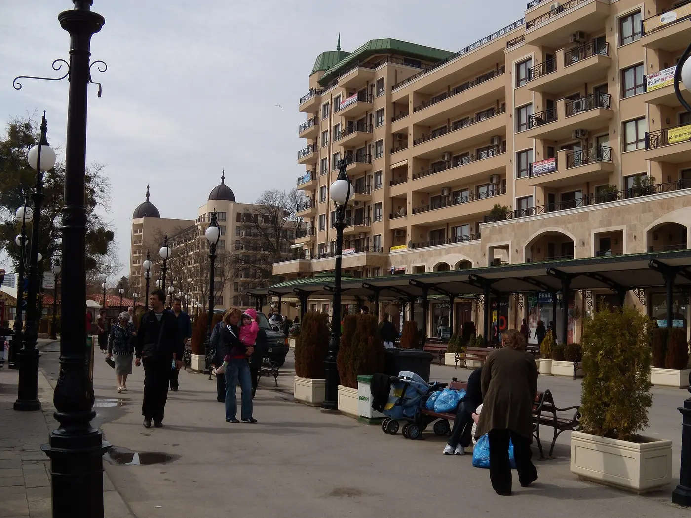 A pedestrian street at Golden Sands lined with large cream resort hotels, black lamp posts and covered shopfronts, people strolling between them