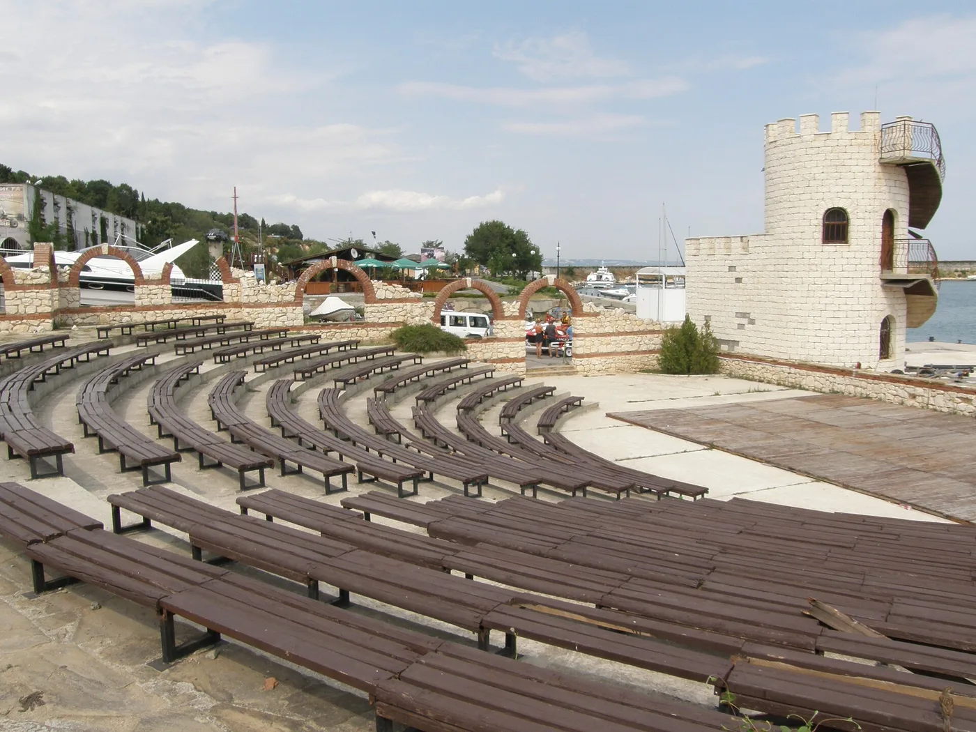 A seafront open-air amphitheatre at Golden Sands with curved rows of wooden benches, a white castle-style tower folly and small marina behind, the sea to the right