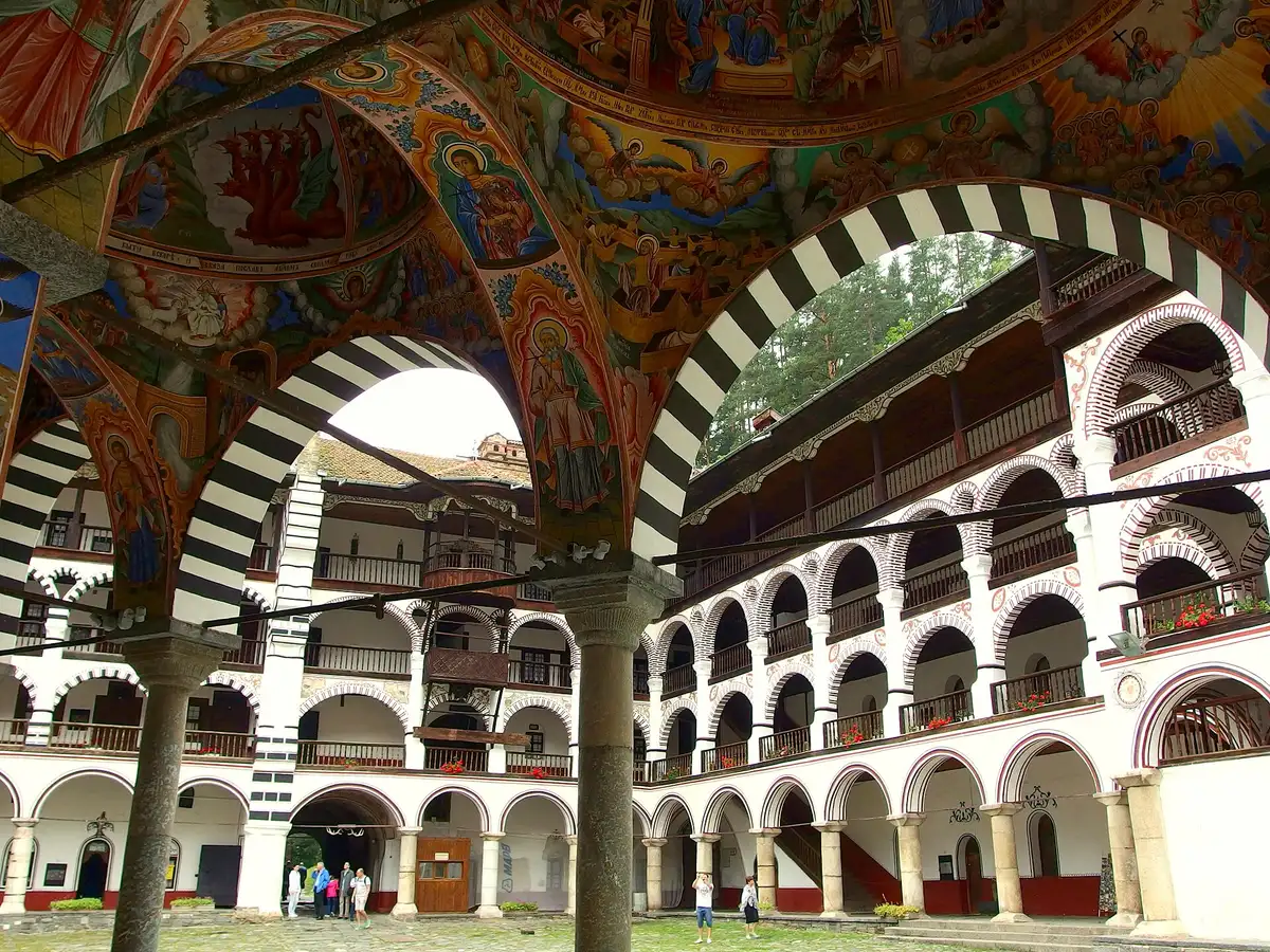 The frescoed portico ceiling and black-and-white striped arches of Rila Monastery, the arcaded courtyard wings rising behind