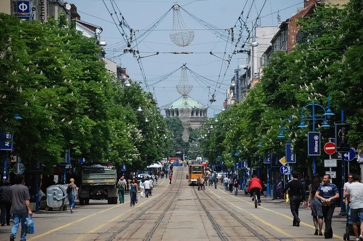 Crowds of people walking along the tree-lined Vitosha Boulevard in central Sofia on a sunny day, with the dome of Sveta Nedelya church in the distance