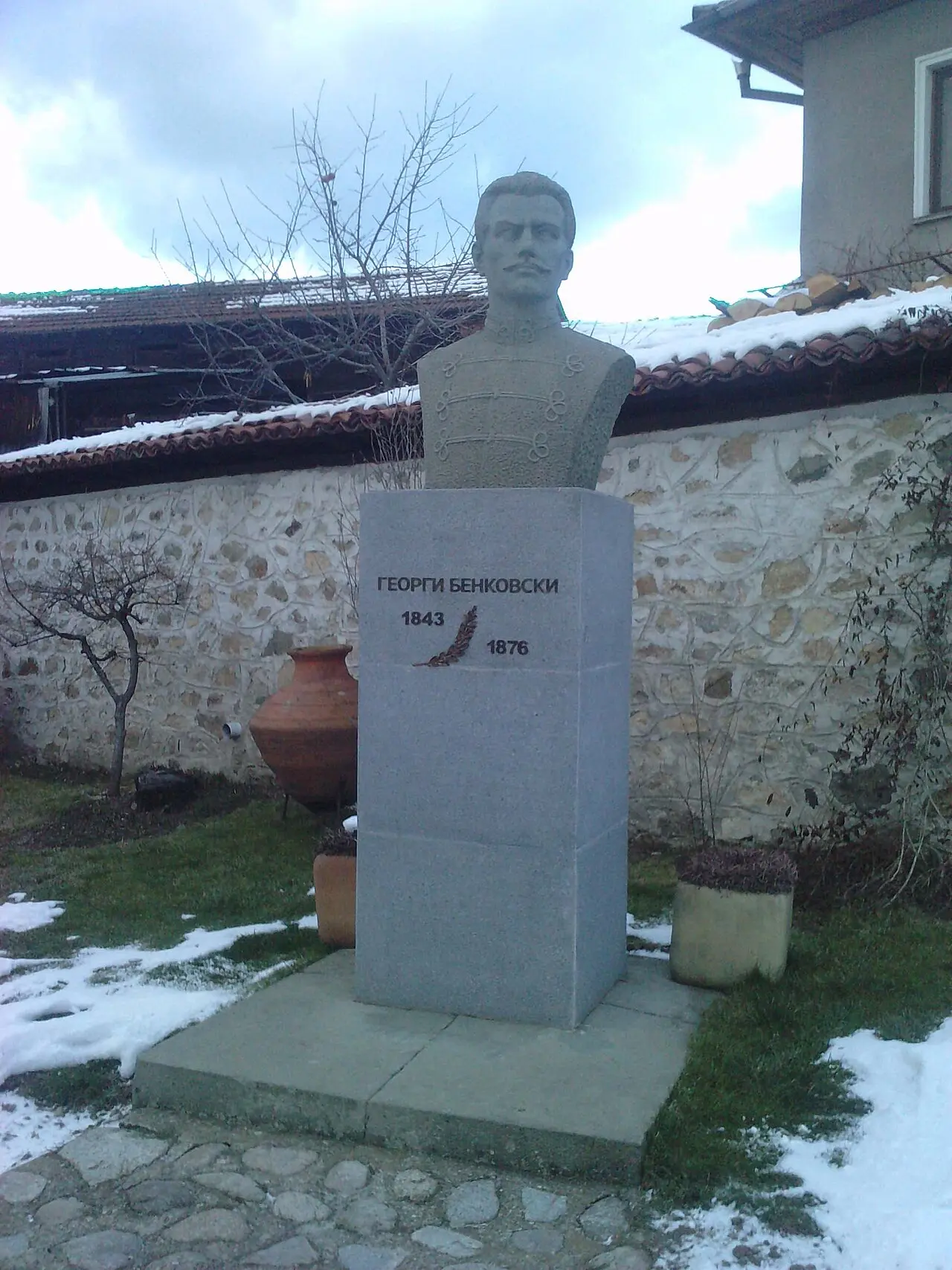 A stone bust of Georgi Benkovski on a plinth reading 1843-1876 with an engraved eagle, in a courtyard by a rubble-stone wall in Koprivshtitsa