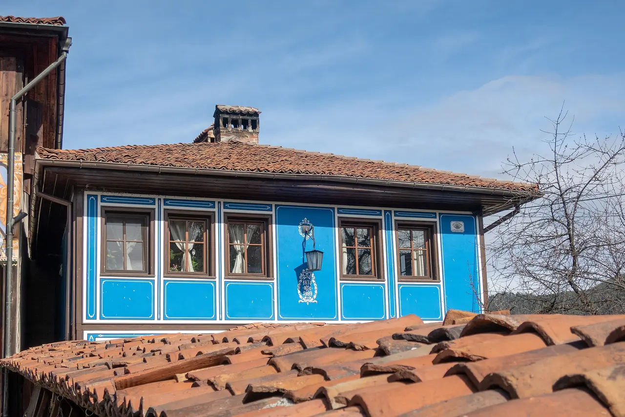 A bright blue Revival house in Koprivshtitsa with white-outlined panels, a hanging wrought-iron lantern and a dated medallion, seen over a foreground of terracotta roof tiles