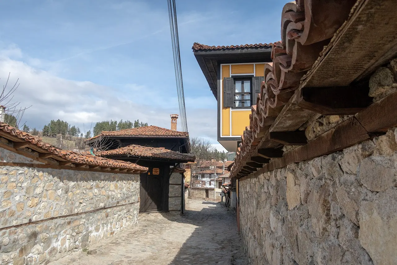 A narrow cobbled lane running between high stone garden walls in Koprivshtitsa, a yellow oriel house leaning overhead and pine-covered hills at the end of the street