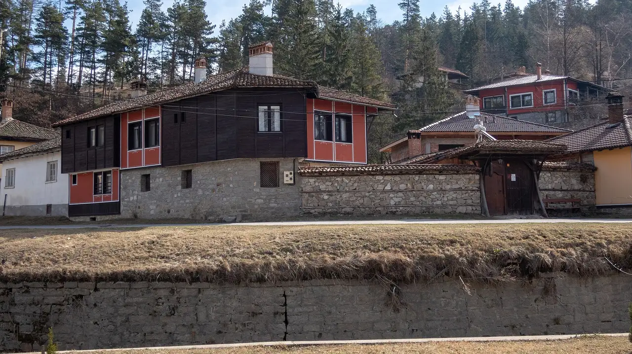 A dark timber and red-panelled National Revival house with an overhanging oriel above a stone wall in Koprivshtitsa, with the forested Sredna Gora hills behind