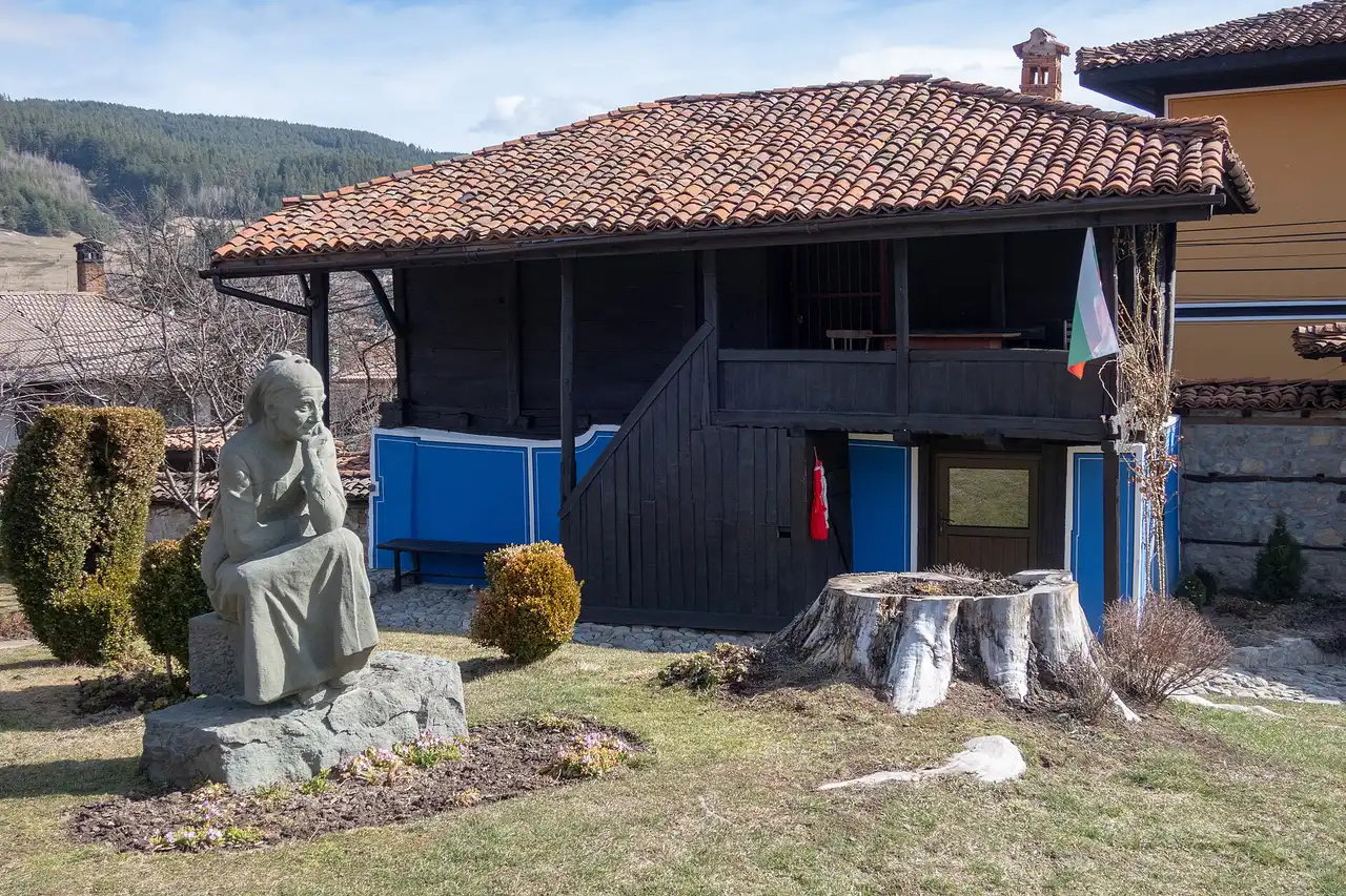 The dark timber Dimcho Debelyanov House in Koprivshtitsa with a seated stone statue of a pensive old woman on a rock in the garden and a Bulgarian flag by the door