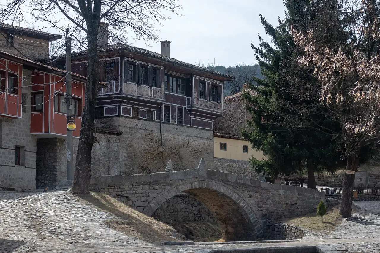A small humpbacked stone bridge over a dry channel on a cobbled square in Koprivshtitsa, with a large timber Revival house behind it