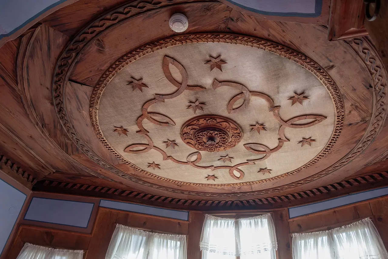 An ornate carved wooden ceiling inside a Koprivshtitsa Revival house: a large sunburst rosette of ribbons and stars radiating from a central boss, with lace-curtained windows below