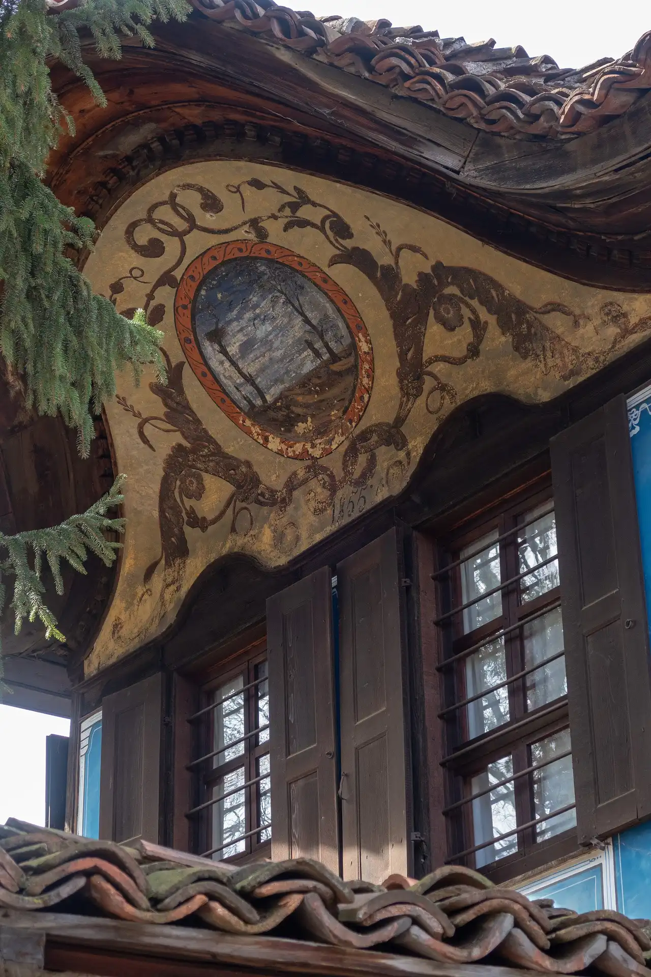 A close-up of a painted alafranga mural on the curved pediment of the Lyutov House in Koprivshtitsa: an oval landscape in an ochre frame surrounded by dark floral scrollwork, dated 1855