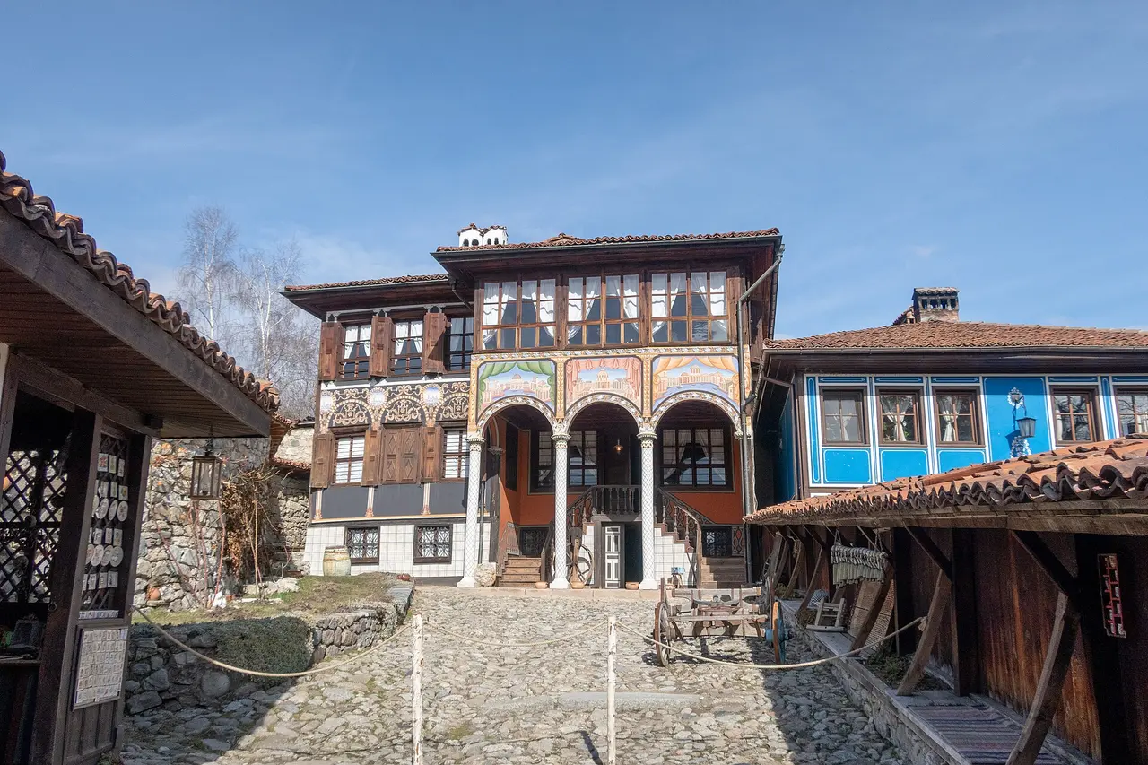 The Oslekov House in Koprivshtitsa: a Revival mansion with a triple-arched painted colonnade and murals over the entrance, a blue-painted house beside it and a cobbled courtyard with a wooden cart