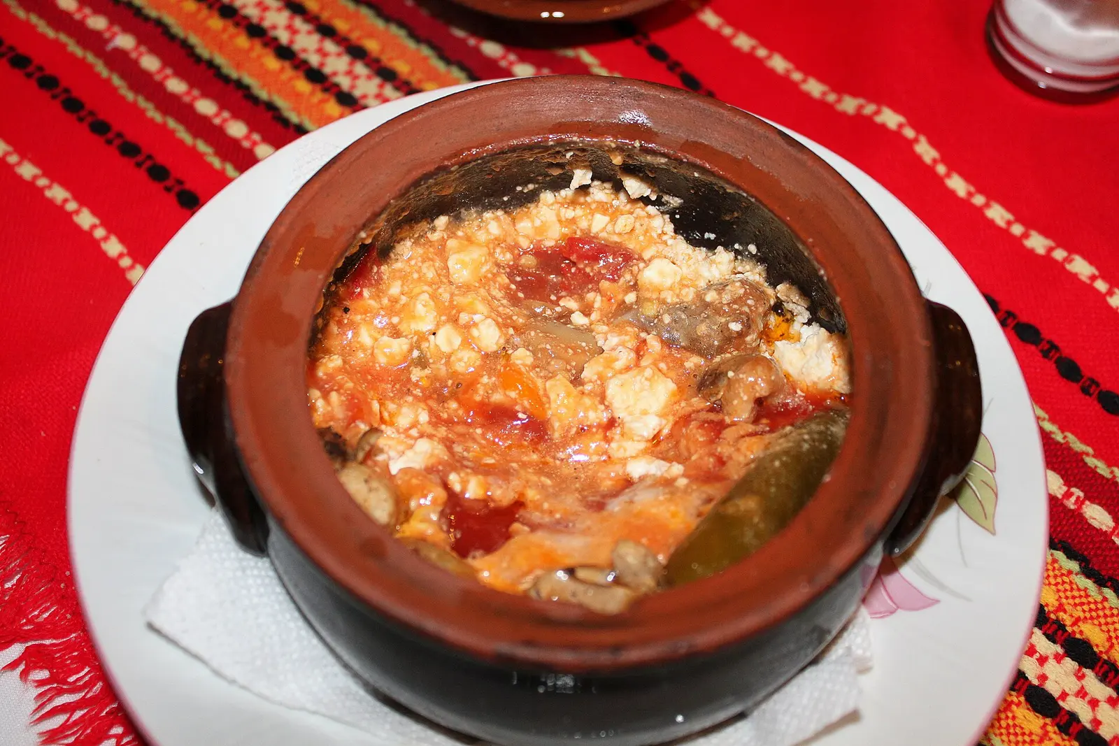 A traditional Bulgarian dish baked and served in a clay pot, topped with melted cheese, on a red folk-pattern tablecloth