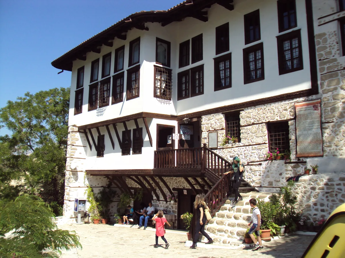 The three-storey white facade of the Kordopulov House in Melnik, with a stone lower floor and a wooden external staircase, visitors on the steps