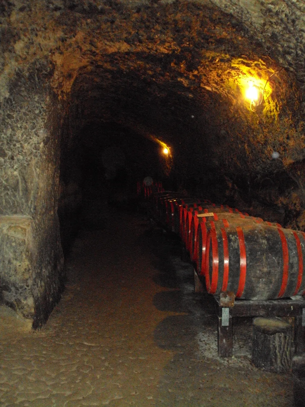 A row of old wooden wine barrels with bright red iron hoops standing along a rock-cut cellar in Melnik, lit by a warm lamp