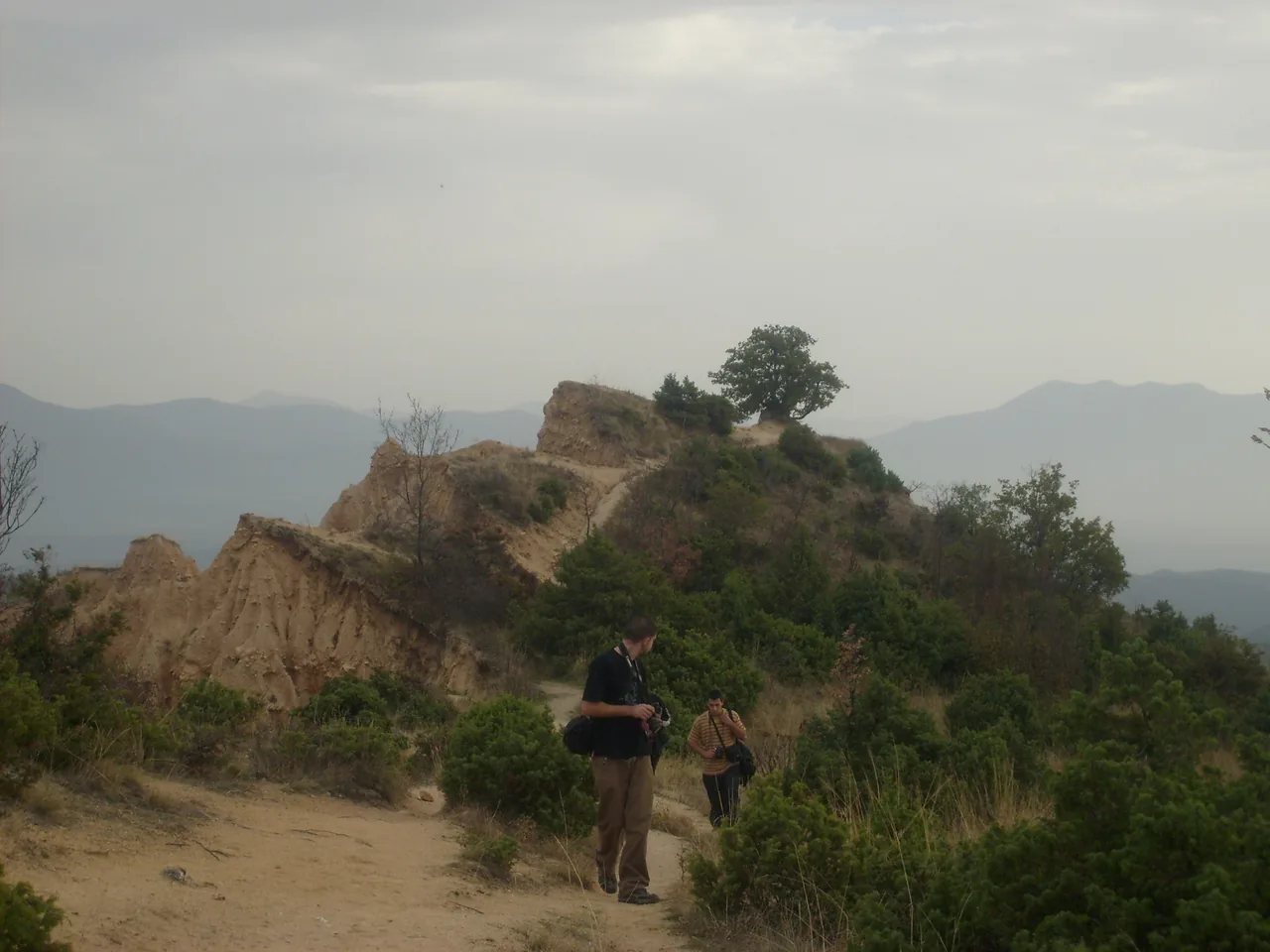 Two walkers on a dirt path along a ridge of sandstone formations near Melnik, a lone tree on the crest