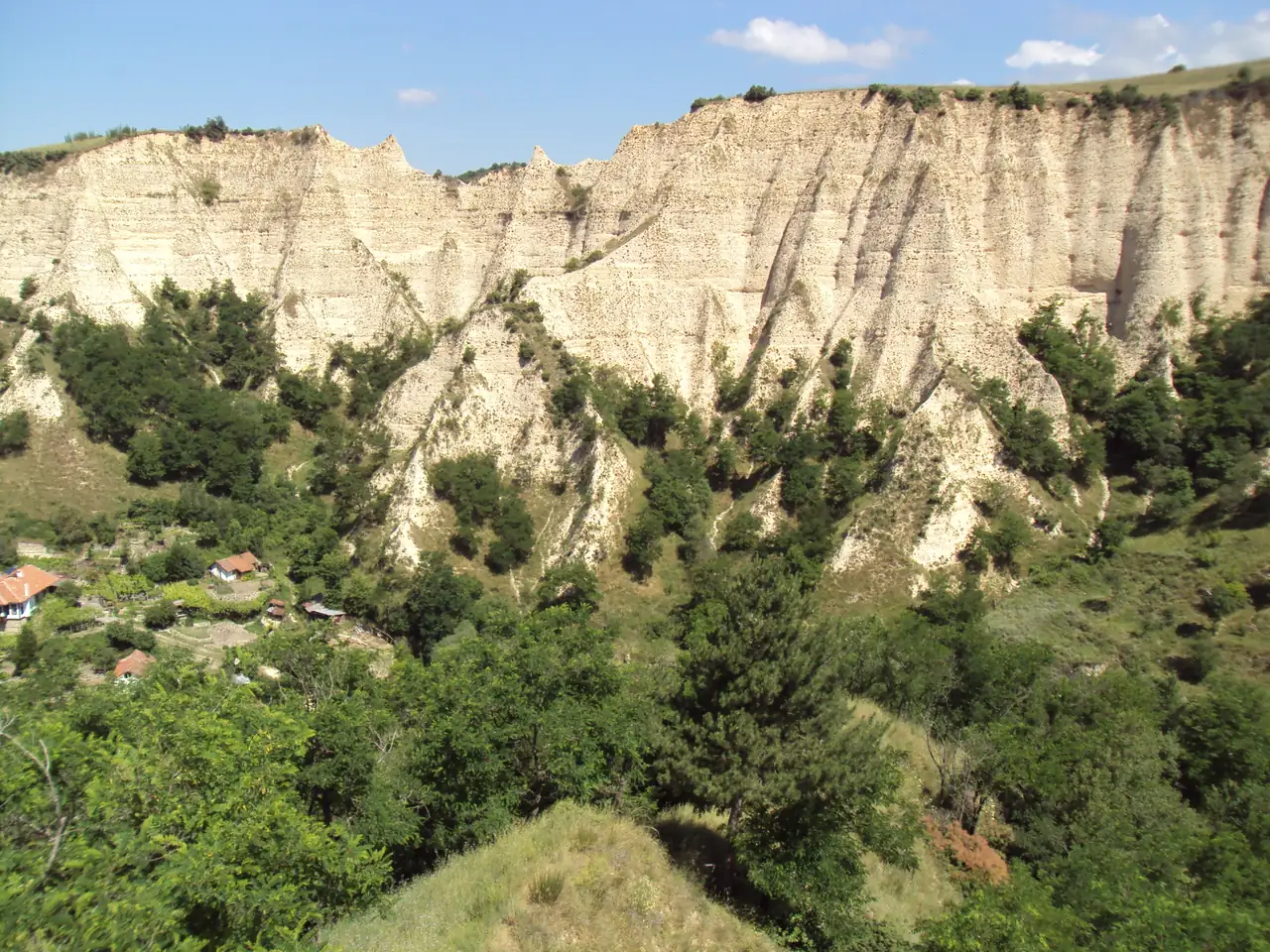 A wall of eroded sandstone pyramids rising above a few red-roofed houses and vineyards on the valley floor near Melnik