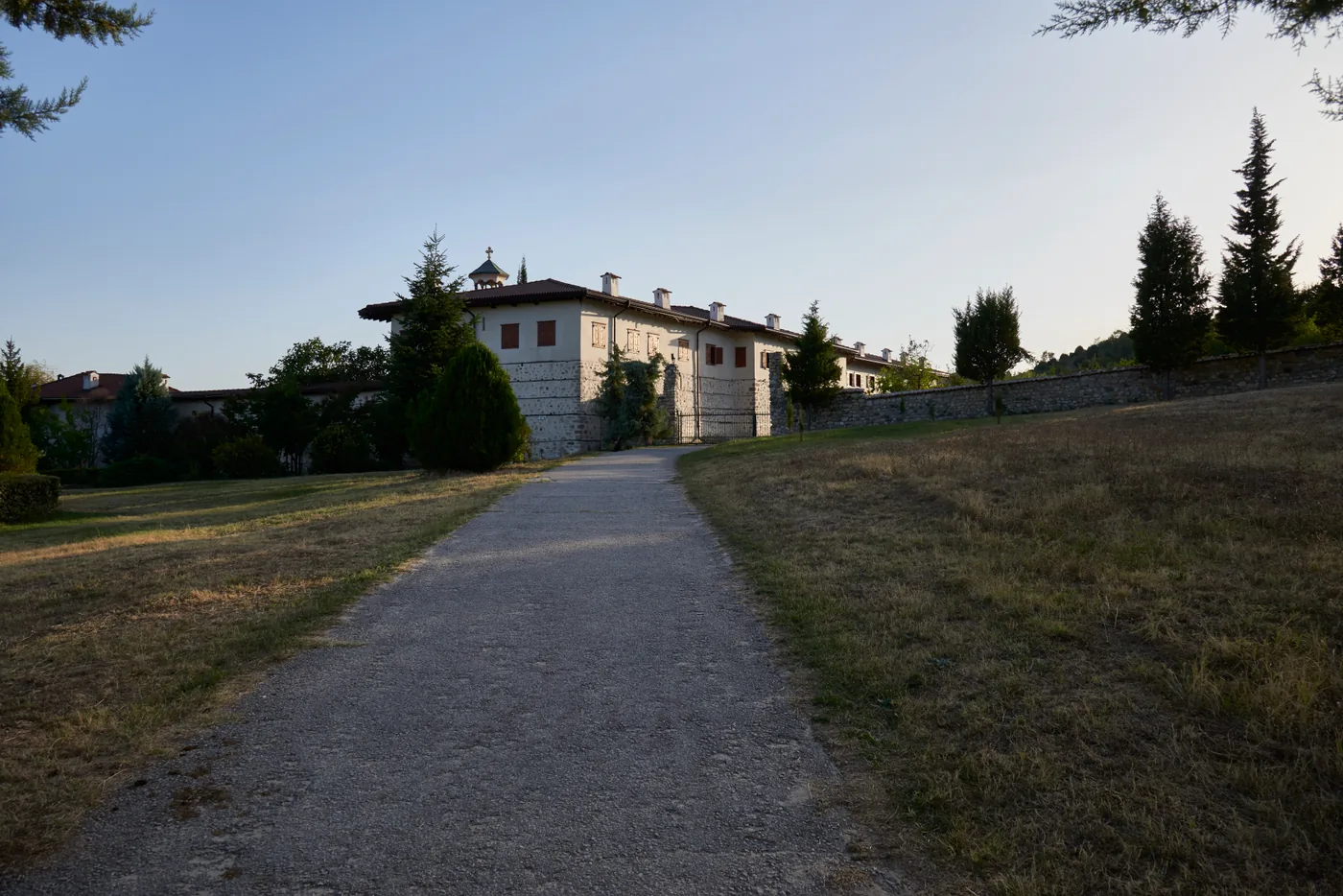 The whitewashed outer walls and tiled roofs of Rozhen Monastery seen from the gravel approach path at dusk
