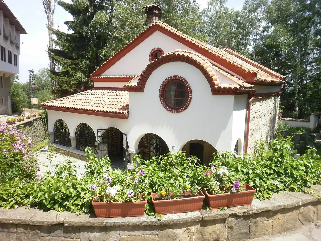 The small white church of Dragalevtsi Monastery with a red tiled roof and arcaded porch, behind flower boxes