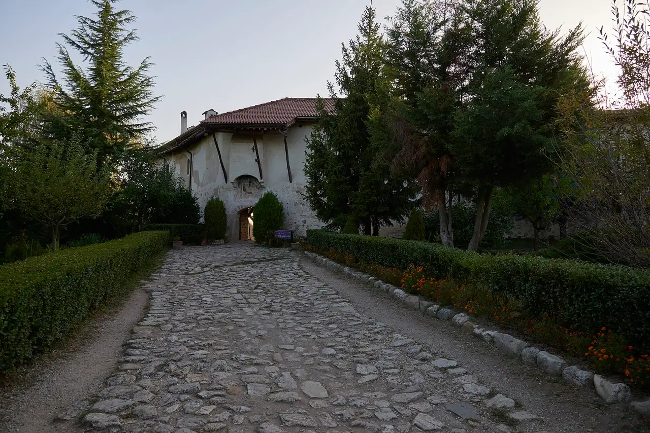 A cobbled path lined with hedges leading up to the gate of Rozhen Monastery at dusk