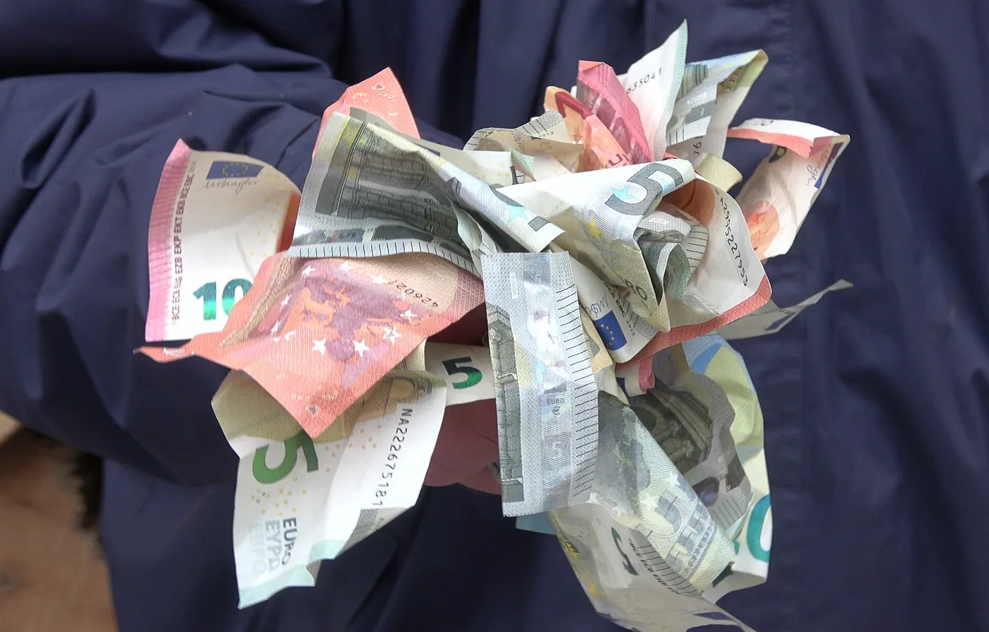 A handful of crumpled euro banknotes - fives, tens, twenties and fifties - held in one hand against a dark jacket