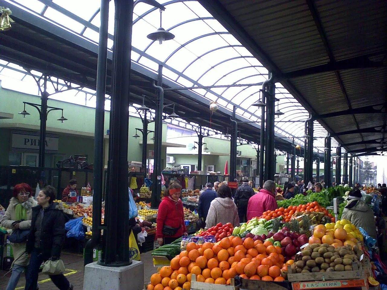 Fruit and vegetable stalls piled with oranges, apples and greens under the glass roof of the covered central market in Burgas, shoppers browsing between the tables