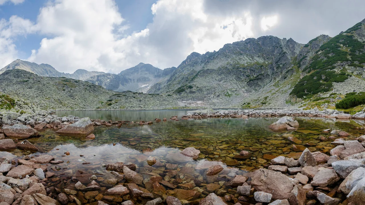 A clear alpine lake in the Rila mountains with the rocky Musala massif rising behind it under a partly cloudy summer sky