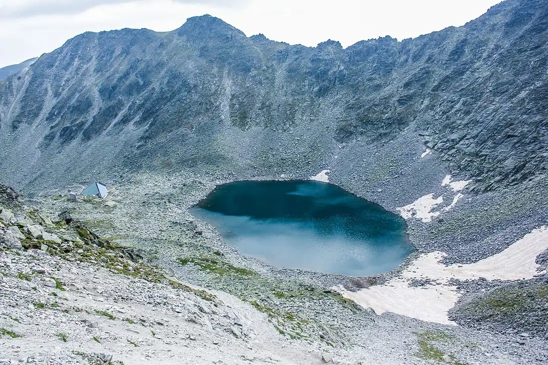 A dark turquoise glacial lake in a rocky cirque in the Rila mountains with patches of snow surviving on its shaded shore