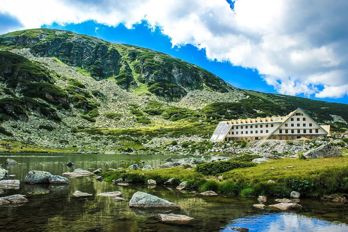 A large A-frame mountain hut beside a small alpine lake in the Rila mountains, with rocky slopes rising behind it