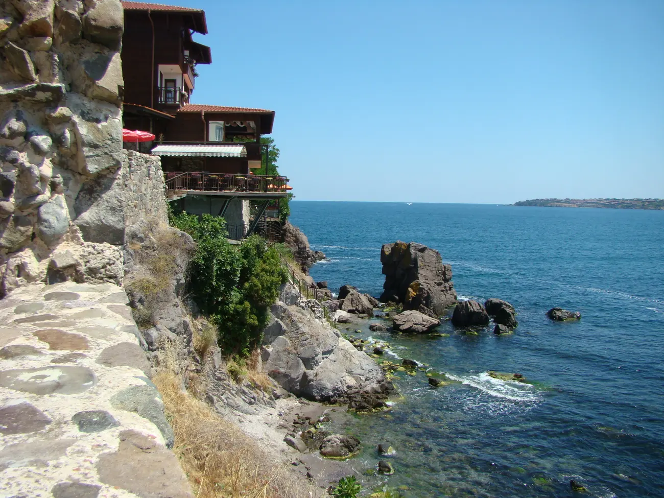 Timber old-town houses in Sozopol perched on the rocks above the sea, a restaurant terrace with a red parasol overlooking a rocky cove