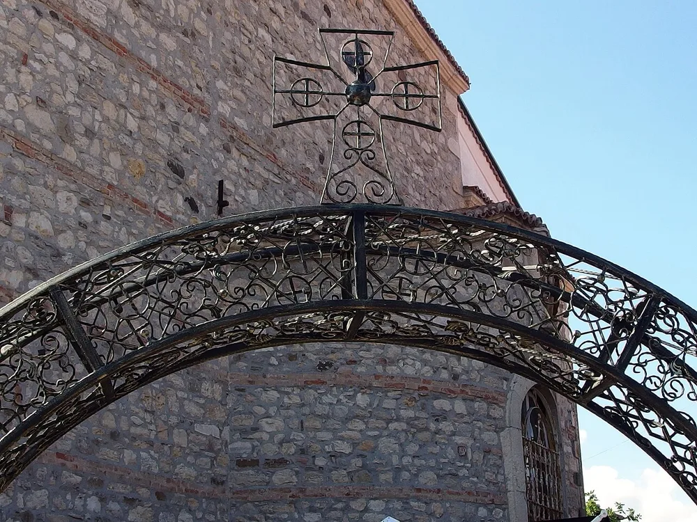 A wrought-iron gate with an ornate forged cross in front of a medieval stone church wall in Nessebar