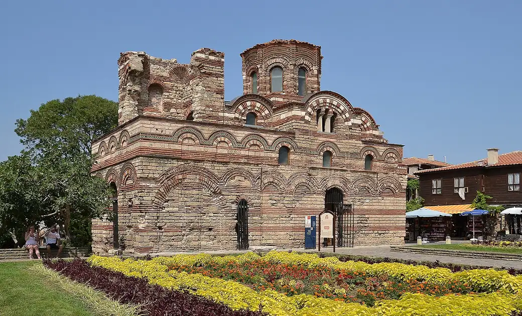The striped brick-and-stone medieval Church of Christ Pantocrator in Nessebar old town, with flower beds and souvenir stalls around it
