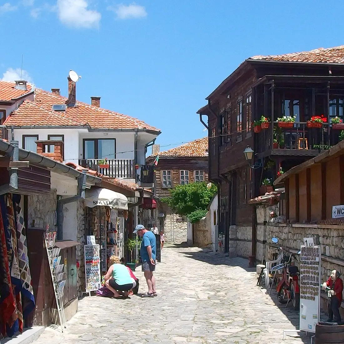 A cobbled lane in Nessebar old town lined with souvenir shops and timber-balconied houses, a few tourists browsing in summer sun