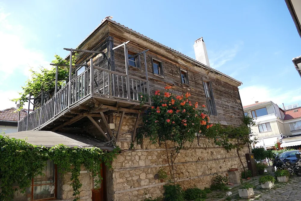 A restored 19th-century Nessebar Revival house with a stone base and an overhanging timber upper floor, roses climbing the wall