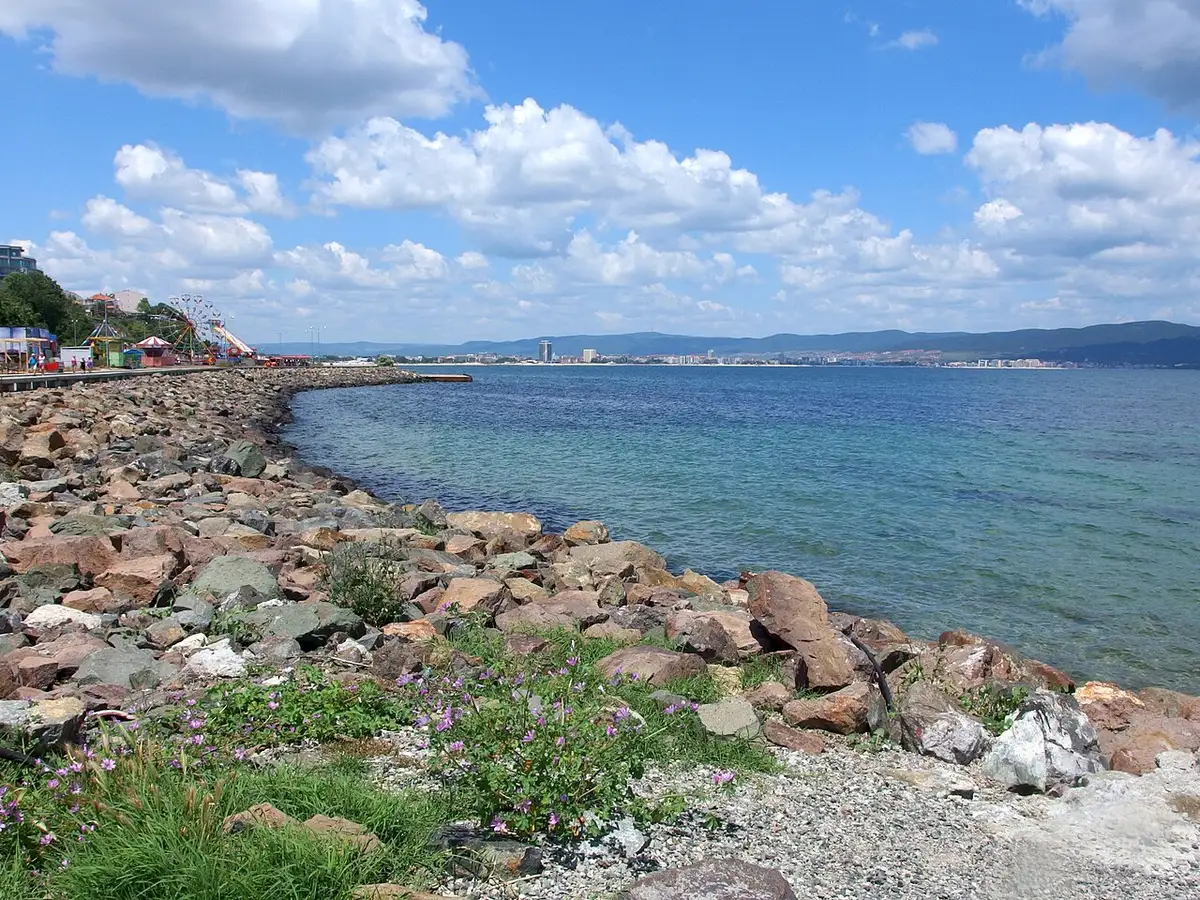 The rocky shore of the Nessebar peninsula looking across the blue bay to Sunny Beach resort under summer clouds