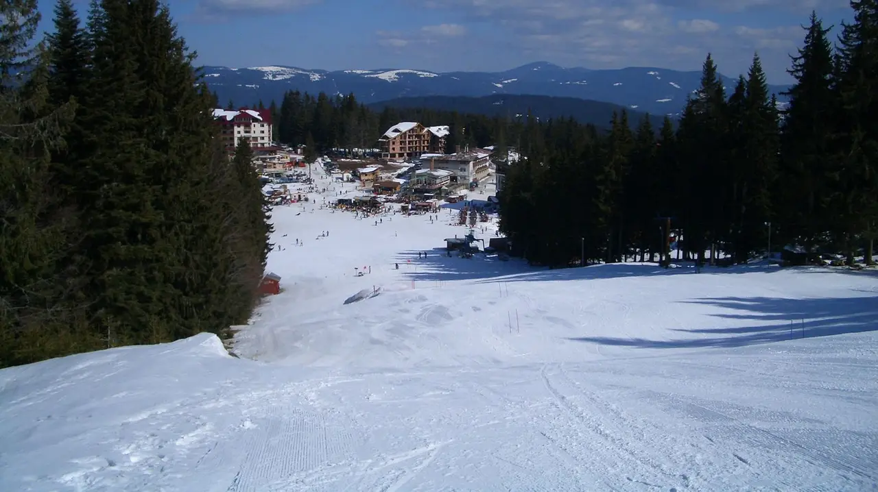 A wide groomed piste running down to the Pamporovo resort base under a blue sky, skiers scattered on the snow with the Rhodope hills behind
