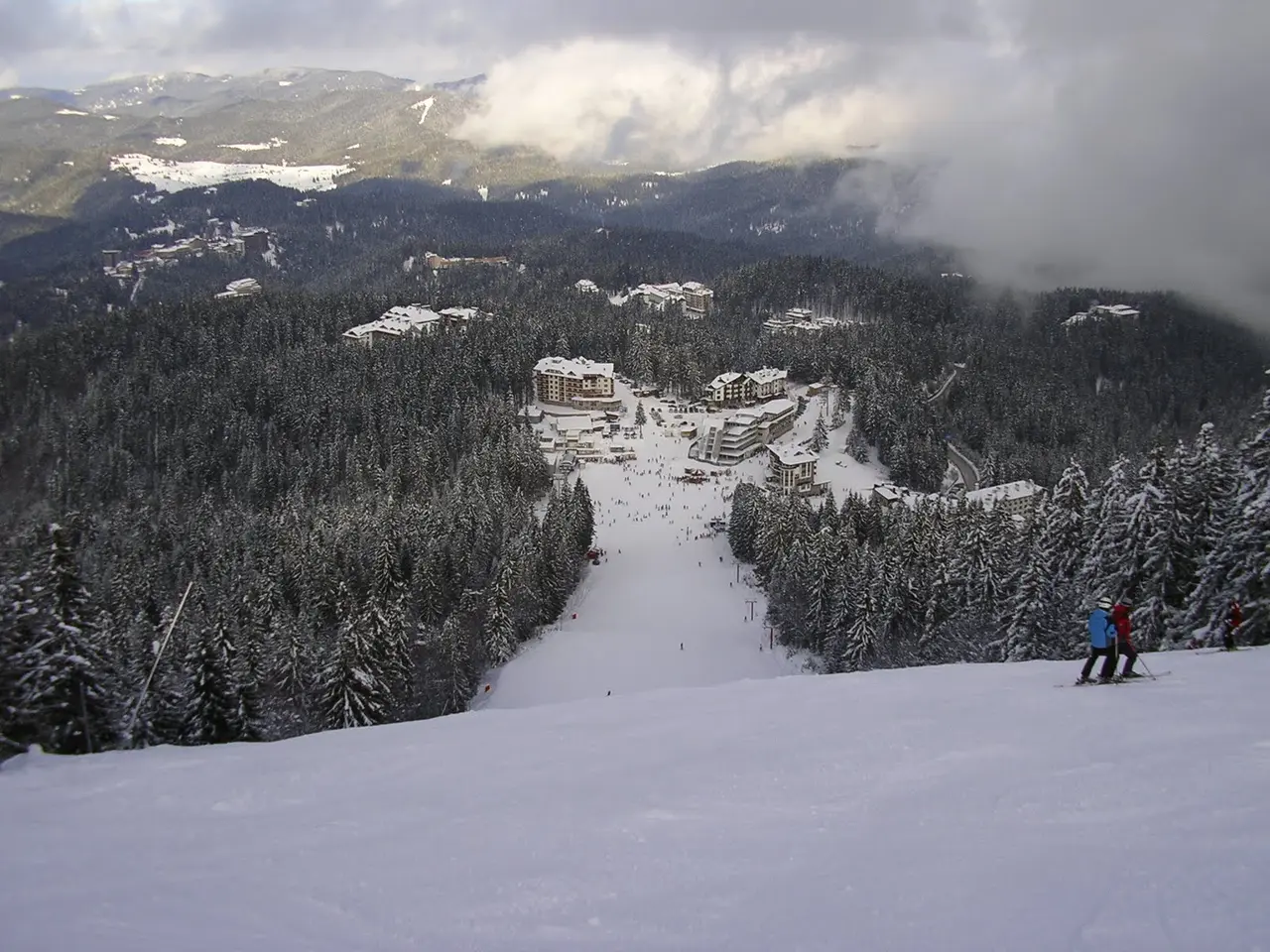 Two skiers descending a wide groomed piste through snowy spruce forest toward the Pamporovo resort base far below