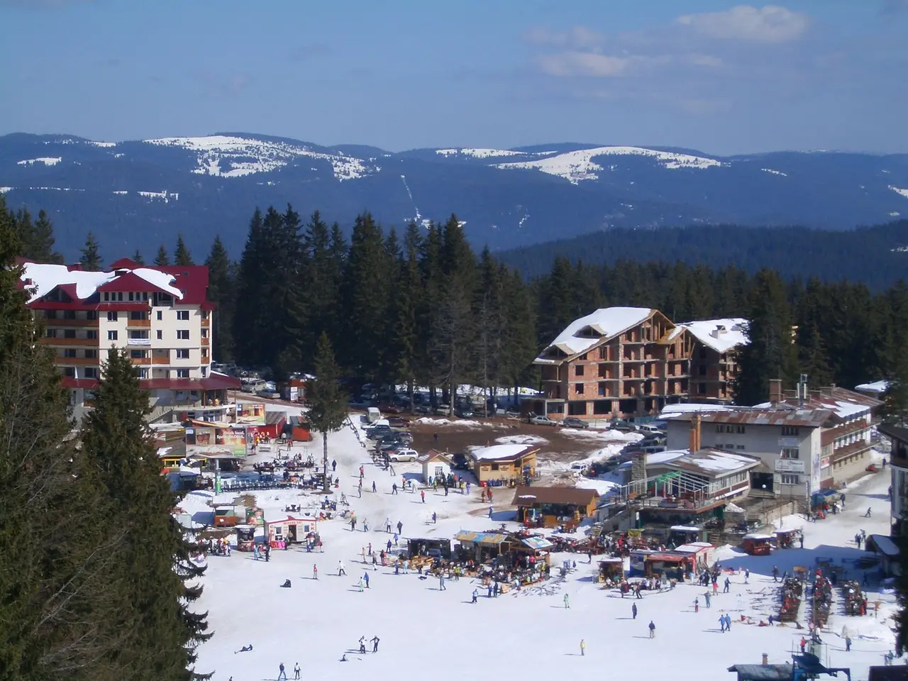 The Pamporovo resort base plaza in winter with skiers, food stalls and hotels ringed by snowy forest and distant Rhodope peaks