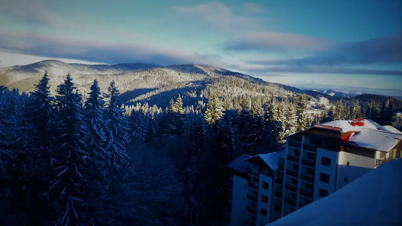A snowy Pamporovo hillside at dusk with frosted spruce forest and a lit apartment hotel on the right