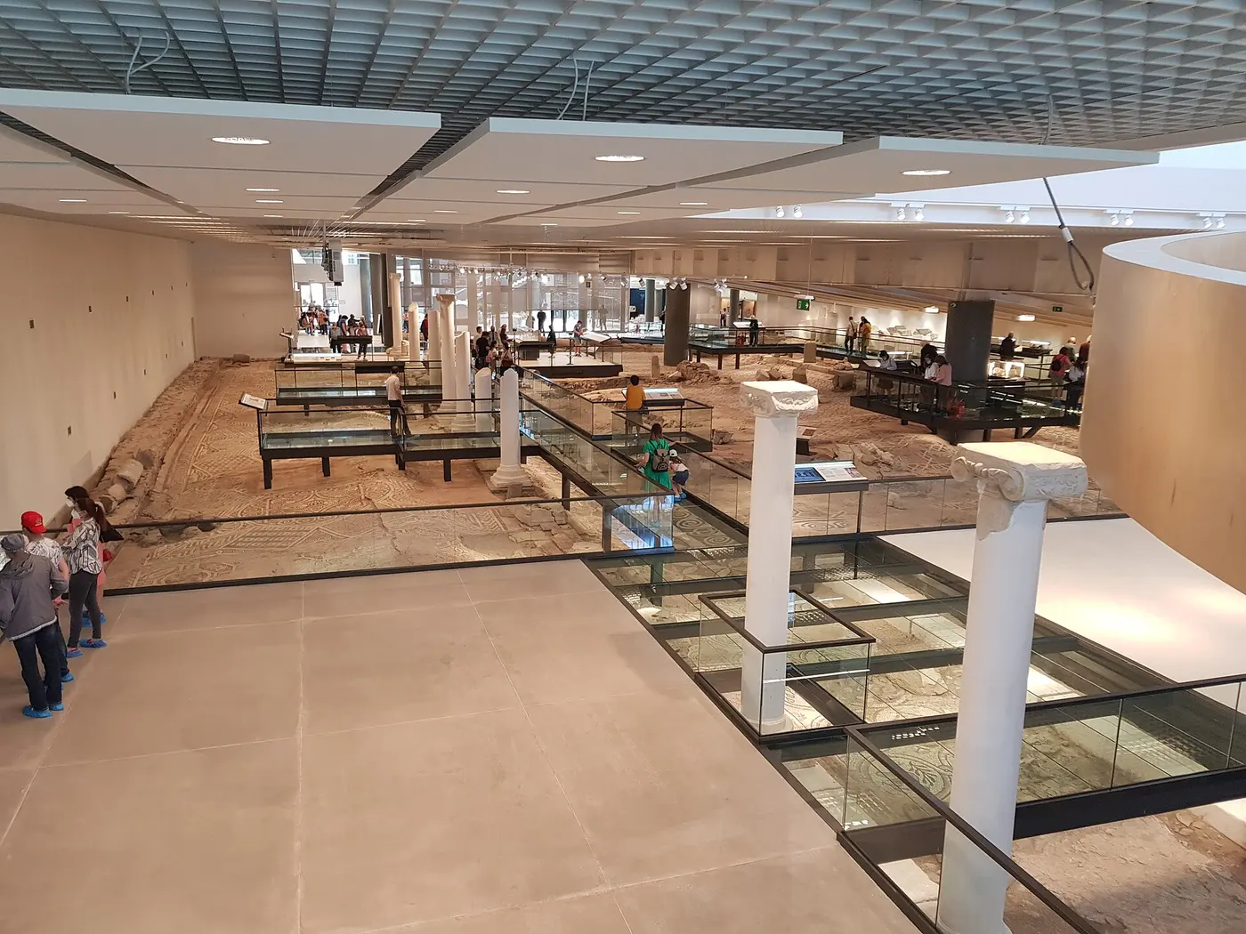 Interior of the Bishop's Basilica museum in Plovdiv, ancient mosaic floors displayed under glass walkways with white columns and visitors