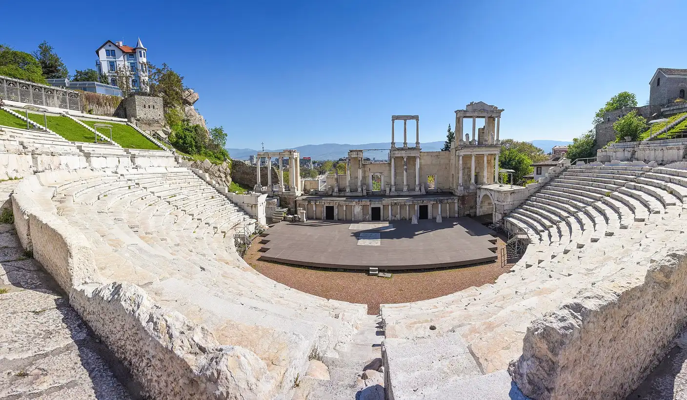 Wide view of the Ancient Roman Theatre of Philippopolis in Plovdiv, marble seating tiers curving around the stage building with its columns, mountains behind